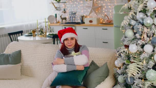 Mujer joven en sombrero de santa con caja de regalo sentada en el sofá cerca del árbol de Navidad en la sala de estar. Feliz hembra considera y abraza presente la Navidad. Concepto de sorpresa durante las vacaciones. — Vídeo de stock