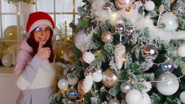 一个大拇指的女人身穿保暖毛衣、头戴圣诞帽的红头发的积极的年轻女性，竖起大拇指，看着站在装饰过的圣诞树旁的相机 — 图库视频影像