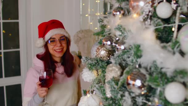 Donna da sogno con vino vicino all'albero di Natale che distoglie lo sguardo. Donna in cappello di Babbo Natale in piedi con bicchiere di vino rosso mentre festeggia il nuovo anno vicino all'albero di Natale decorato a casa. — Video Stock