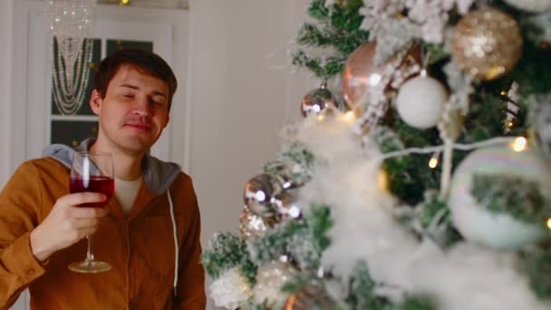 Un uomo che beve vino vicino all'albero di Natale. Maschio bere vino rosso dal vetro durante la celebrazione del nuovo anno vicino all'albero di Natale decorato a casa — Video Stock