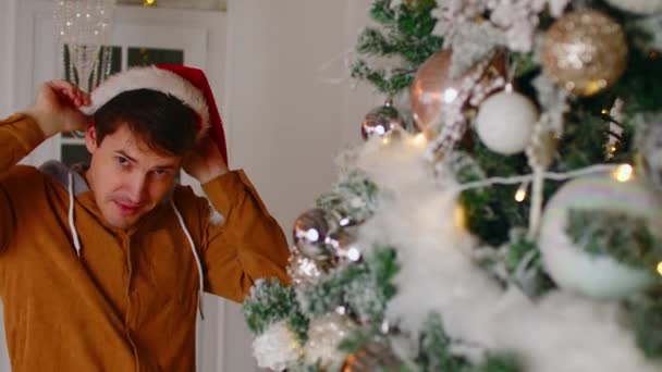 Позитивный человек, стоящий рядом с елкой. Молодой мужчина носит шляпу Санты. Счастливый мужчина смотрит в камеру и улыбается, празднуя Новый год у украшенной елки дома. — стоковое видео
