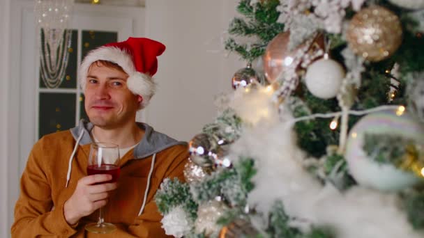 Uomo da sogno con vino vicino all'albero di Natale che distoglie lo sguardo. Maschio in cappello di Babbo Natale in piedi con bicchiere di vino rosso mentre festeggia il nuovo anno vicino all'albero di Natale decorato a casa. — Video Stock