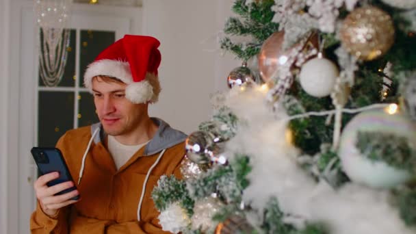Людина використовує смартфон під час Різдва. Чоловік у повсякденному одязі та капелюсі Санта стоїть біля ялинки та переглядає мобільний телефон вдома — стокове відео