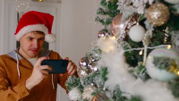 Мужчина фотографирует ёлку. Молодой мужчина в шляпе Санта-Клауса фотографируется на мобильный телефон новогодней елки, украшенной сказочными огнями и безделушками во время новогодних праздников — стоковое видео