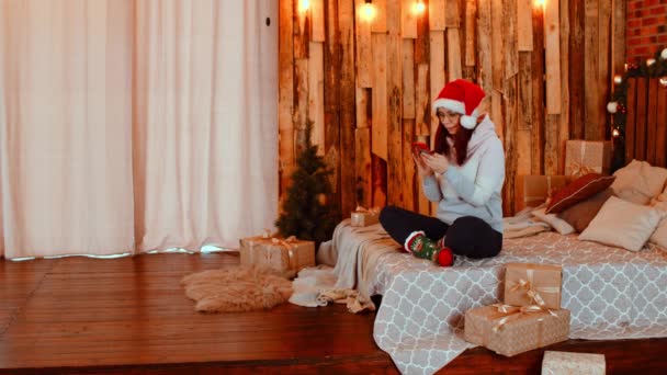 Молодая женщина в шляпе Санта-Клауса просматривает смартфон, сидя на кровати. Очаровательная женщина отдыхает с мобильным телефоном и празднует Рождество дома. Концепция праздников и хорошее настроение. — стоковое видео
