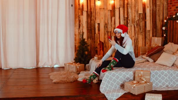 Młoda kobieta w czapce Świętego Mikołaja przegląda smartfona, siedzi na łóżku. Urocza kobieta odpoczywa z telefonem komórkowym i świętuje święta w domu. Pojęcie wakacji i dobry nastrój. — Wideo stockowe