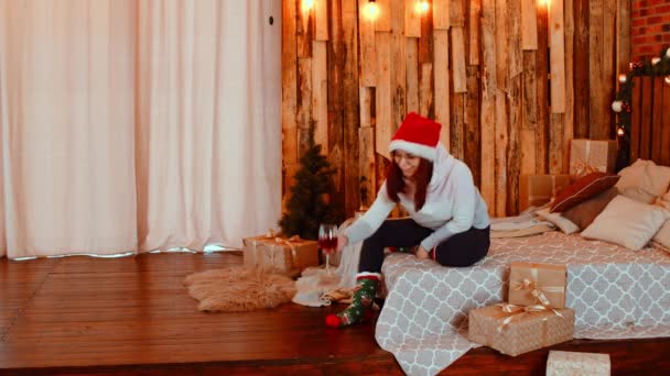 Young woman in santa hat and glasses sitting on bed and drinking red wine. Positive female resting with alcohol and celebrating New Year. Woman sitting surrounded by gift boxes and Christmas tree. — Stock Video