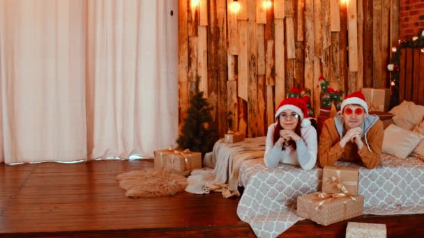 Homem e mulher em chapéus de Papai Noel e óculos deitados na cama cercados por caixas de presente e árvore de Natal durante as festas. Casal feliz olhando para a câmera, sorrindo e balançando as pernas. — Vídeo de Stock