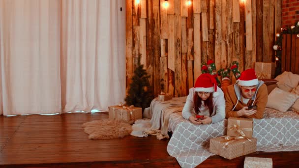 Couple dans Santa chapeaux accro aux médias sociaux et les téléphones cellulaires couchés sur le lit parmi les cadeaux présentés attachés avec des rubans en studio avec des lampes lumineuses et Noël — Video