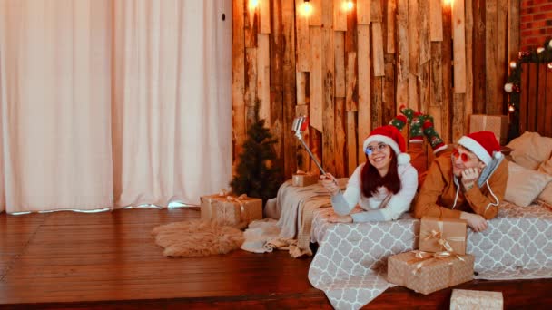 Positiva pareja joven en los sombreros de Santa acostado en la cama cómoda y tomar selfie en el teléfono inteligente conectado en palo durante las vacaciones de Navidad entre los regalos envueltos en el acogedor dormitorio — Vídeo de stock