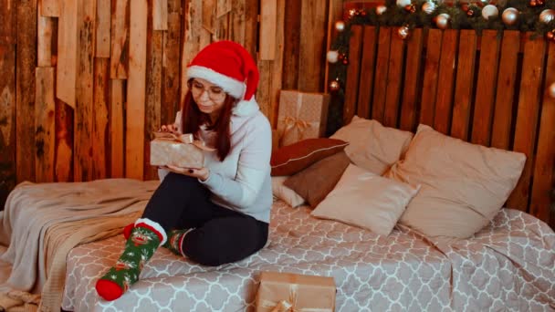 Smiling woman with Christmas gift. Young smiling female in Santa hat warm clothes and eyeglasses sitting on bed with wrapped Christmas gift box in studio — Stock Video