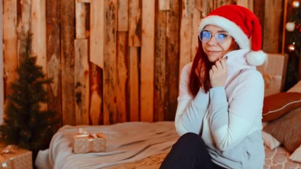 Mulher em casa durante o Natal. Jovem feliz em roupas quentes e chapéu de Papai Noel sorrindo e olhando para a câmera enquanto sentado em cama confortável em casa de campo de madeira aconchegante durante as férias de Natal — Vídeo de Stock