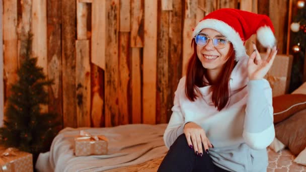 Wanita muda bertopi santa dan kacamata bermain dengan pompom, melihat ke kamera. Wanita cantik duduk di tempat tidur dan bersenang-senang dengan topi Natal. — Stok Video