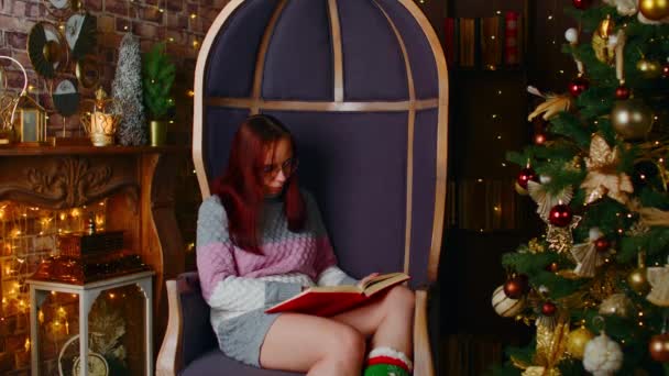 暖かいセーターと眼鏡読書本の集中女性クリスマスツリーの近くの空き時間に居心地の良いアパートで手袋を着用して装飾 — ストック動画