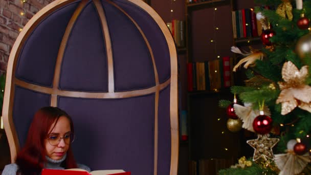 Сконцентрированная женщина в теплых свитерах и очках читает книгу в свободное время возле елки, украшенной перчаточными гирляндами в уютной квартире — стоковое видео