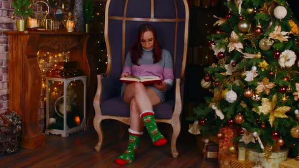 Geconcentreerde vrouw in warme trui en bril lezen boek in vrije tijd in de buurt van kerstboom versierd met gloeiende bloemenslingers in gezellig appartement — Stockvideo