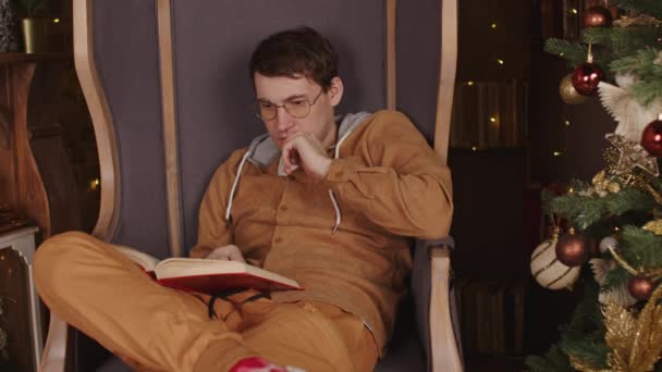 Мужчина читает книгу возле елки. Сконцентрированный мужчина в теплом свитере и очках читает книгу в свободное время возле елки в уютной квартире. — стоковое видео