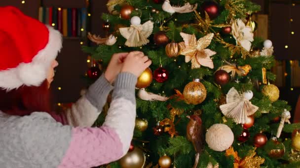 Вид сбоку женщины в шляпе санта, украшающей елку. Женщина в рождественской шляпе вешает рождественскую игрушку и создает праздничную атмосферу во время праздников в помещении. — стоковое видео