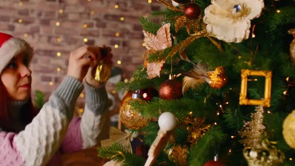 Pemandangan samping wanita dengan topi santa yang menghias pohon Natal. Perempuan dalam topi Natal menggantung mainan Natal dan menciptakan suasana meriah selama liburan di dalam ruangan. — Stok Video