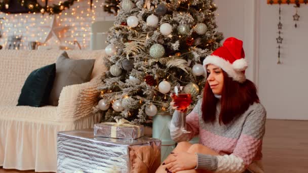 Giovane donna in cappello da Babbo Natale seduta sul pavimento circondata da scatole regalo e albero di Natale. Positivo alcol femminile e festeggiare il nuovo anno in atmosfera festiva. — Video Stock