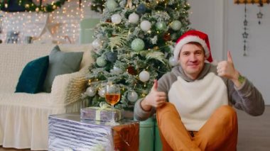 Noel Baba şapkalı genç adam hediye kutuları ve Noel ağacı ile çevrili yerde oturuyor. Pozitif erkek, festival atmosferinde yeni yılı kutluyor..