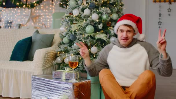 ギフトボックスやクリスマスツリーの近くの床に座ってサンタの帽子の若い男性。正の男は踊り、お祝いの雰囲気の中で新年を祝いながら平和のジェスチャーを示す. — ストック動画