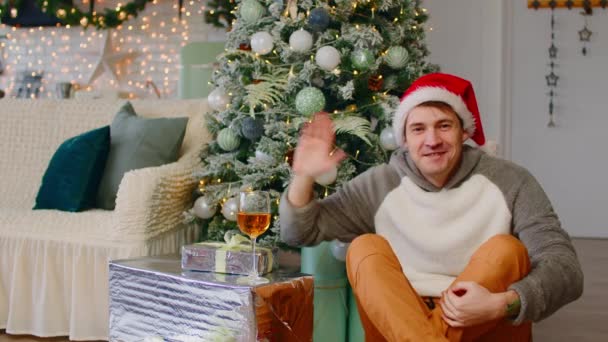 Młody człowiek w kapeluszu Mikołaja siedzi na podłodze otoczony pudełkami z prezentami i choinką. Pozytywny mężczyzna macha ręką, świętując Nowy Rok w świątecznej atmosferze. — Wideo stockowe