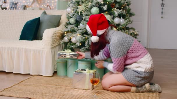 Giovane donna in cappello di Babbo Natale e maglione lavorato a maglia seduta sul pavimento vicino all'albero di Natale. La femmina felice mette regali sotto albero decorato e beve lo champagne, festeggiando vacanze. Concetto di sorpresa. — Video Stock