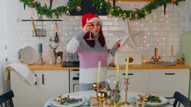Młoda kobieta w noworocznym kapeluszu pije wino i tańczy przy stole. Zbliżenie dorosłych kobiet zabawiających się lampką wina w kuchni. — Wideo stockowe