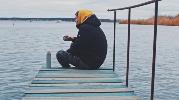 Man on pier drinking tea. Side view of young male in warm clothes sitting on wooden pier of rippling river and enjoying hot drink from thermos while admiring nature in cold autumn day — Stockvideo