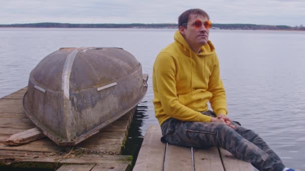 Młody człowiek w jasnych okularach i bluzie siedzi na drewnianym molo i rozgląda się na tle płynącej wody. Zrelaksowany mężczyzna spoczywa na molo w pobliżu łodzi w zimną pogodę. — Wideo stockowe