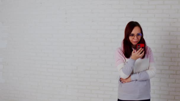 Mujer joven usando teléfono inteligente cerca de la pared. Mujer joven con el pelo largo en sudadera con capucha rosa y gafas de mensajería en el teléfono móvil mientras está de pie contra la pared de ladrillo blanco — Vídeo de stock