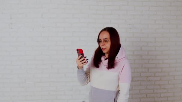 Mujer joven usando teléfono inteligente cerca de la pared. Mujer joven con el pelo largo en sudadera con capucha rosa y gafas de mensajería en el teléfono móvil mientras está de pie contra la pared de ladrillo blanco — Vídeo de stock