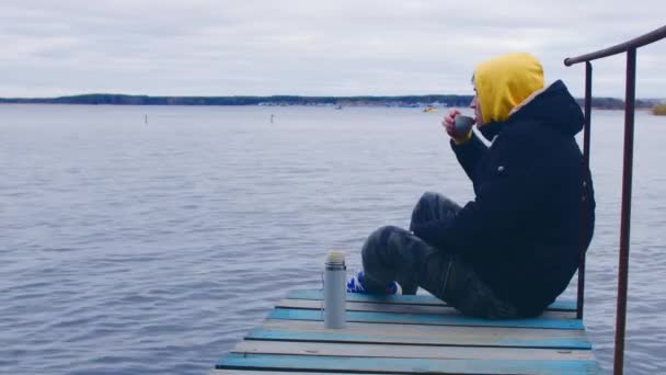 Человек на пирсе пьет чай. Боковой вид молодого мужчины в теплой одежде, сидящего на деревянном пирсе бурлящей реки и наслаждающегося горячим напитком из термоса, любуясь природой в холодный осенний день — стоковое видео