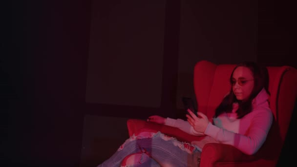 Kobieta pisząca na smartfonie na fotelu. Widok z boku spokojnej młodej kobiety w luźnych ubraniach i okularach spoczywających na wygodnym fotelu i przy użyciu smartfona w ciemnym pokoju z czerwonym oświetleniem — Wideo stockowe