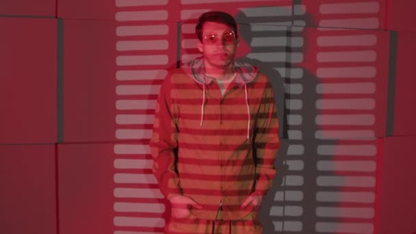 Mężczyzna pod ścianą z cieniami jalousie. Poważny mężczyzna w okularach i ubraniach casual stojący w pobliżu szklanej ściany i ściany z cieniami jalousie w pokoju z czerwonym światłem — Wideo stockowe