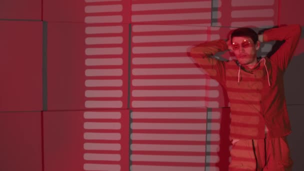 Man in de buurt van muur met schaduwen van jaloezie. Serieuze man in bril en casual kleding staan in de buurt van glazen muur en muur met schaduwen van jaloezie in de kamer met rood licht — Stockvideo