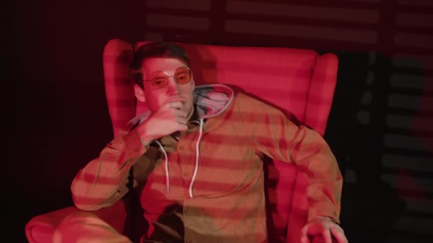 Przemyślany człowiek siedzący w fotelu w ciemnym pokoju z padającym czerwonym światłem i żaluzjami. Zamyślony mężczyzna w okularach drapiący podbródek, odwracając wzrok od życia. — Wideo stockowe
