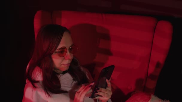 Kobieta pisząca na smartfonie na fotelu. Widok z boku spokojnej młodej kobiety w luźnych ubraniach i okularach spoczywających na wygodnym fotelu i przy użyciu smartfona w ciemnym pokoju z czerwonym oświetleniem — Wideo stockowe
