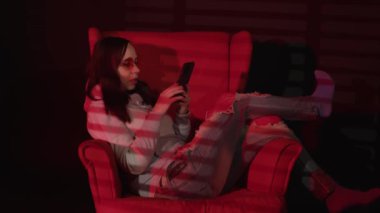 Koltukta akıllı telefondan mesaj atan bir kadın. Rahat koltukta dinlenen rahat giysili ve gözlüklü sakin genç bayanın yan görüntüsü ve kırmızı ışıklı karanlık odada akıllı telefon kullanımı.