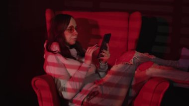 Koltukta akıllı telefondan mesaj atan bir kadın. Rahat koltukta dinlenen rahat giysili ve gözlüklü sakin genç bayanın yan görüntüsü ve kırmızı ışıklı karanlık odada akıllı telefon kullanımı.