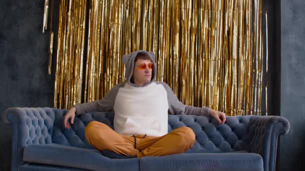 Young man in hood with ears and bright glasses sitting on sofa against wall with golden tinsel. Relaxed male sitting with his feet on couch leaning against back. Concept of rest and free time. — Stockvideo
