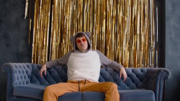 Serious and dissatisfied man in hood with ears and bright glasses sitting on sofa against wall with golden tinsel. Relaxed important male sprawled on couch and looking at camera. — Video Stock