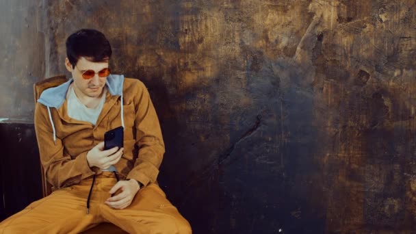 Стильний чоловік у яскравих окулярах сидить на стільці, переглядаючи мобільний телефон біля похмуро-коричневої стіни. Цікавий чоловік використовує смартфон на коричневому фоні з місцем для тексту . — стокове відео