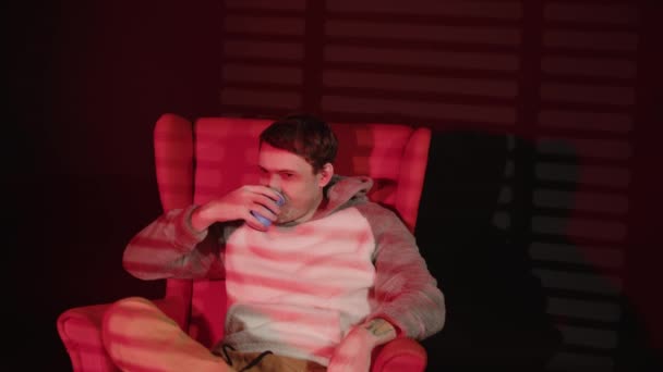 Молодий чоловік з паперовою чашкою кави сидить у кріслі з падаючим світлом з вікна через жалюзі. Розслаблений чоловік п'є каву в темній кімнаті . — стокове відео