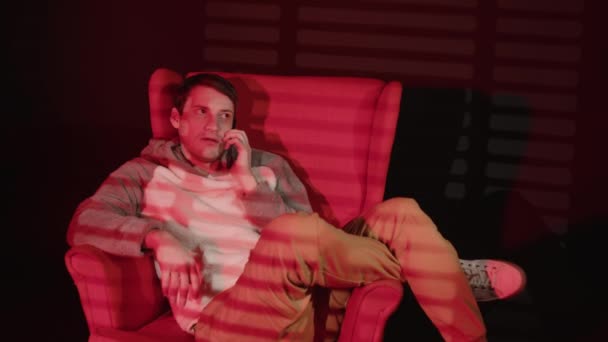 젊은 남자가 스마트폰으로 말을 하고, 어두운 방의 안락 의자에 앉아 붉은 빛 과어두운 블라인드와 함께 있습니다. 분주 한 남자 휴대 전화로 연설하는 모습. — 비디오