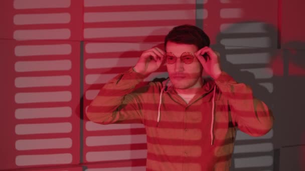 En mann tar på seg briller og står ved veggen med skygger av jalousie. Stilig mann i briller som ser bort i mørke rom med rødt lys og skyggepersienner. – stockvideo