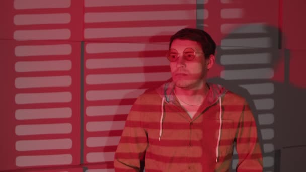 Omul de lângă perete cu umbre de jalousie. Bărbat serios în ochelari și haine casual în picioare lângă perete de sticlă și perete cu umbre de jalousie în cameră cu lumină roșie — Videoclip de stoc
