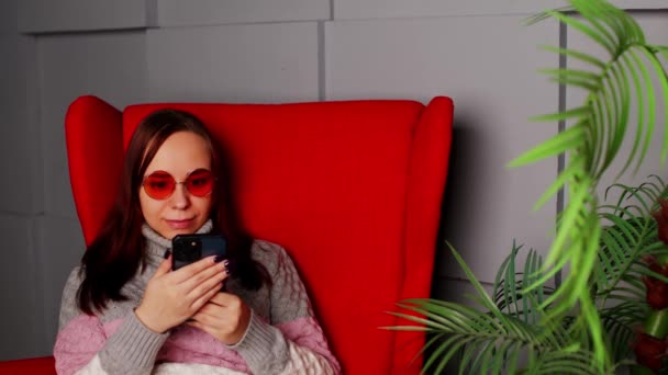 Kobieta w stylowych okularach słonecznych siedzi w fotelu ze smartfonem. Poważna młoda kobieta z ciemnymi włosami w luźnych ubraniach siedzi w fotelu i wysyła wiadomości na smartfonie w domu — Wideo stockowe