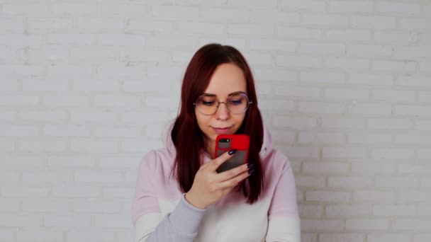 Jonge vrouw met smartphone in de buurt van muur. jong wijfje met lang haar in roze hoodie en bril messaging op mobiele telefoon terwijl staan tegen witte baksteen muur — Stockvideo